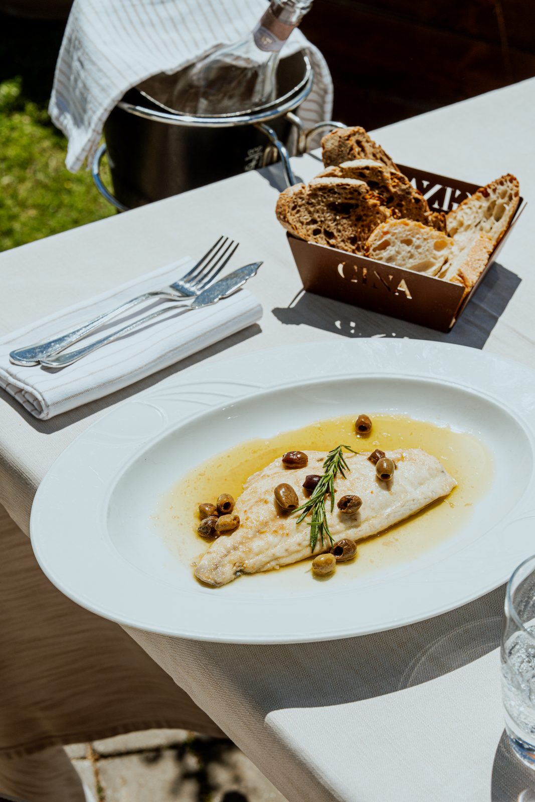 Un filetto di pesce con le olive e un rametto di rosmarino, servito a un tavolo all'aperto accanto al cestino del pane