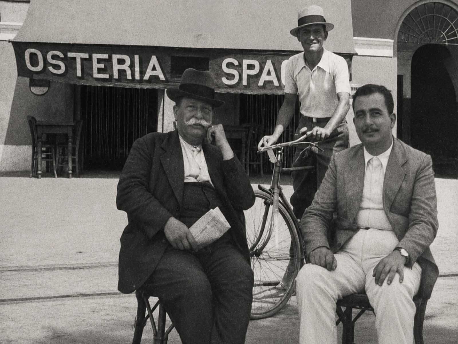 Fotografia d'archivio in bianco e nero degli anni '50: l'esterno dell'Osteria La Spacca, con la tenda-insegna «OSTERIA SPACCA» sopra l'ingresso e i tavolini all'interno. Davanti al locale, in strada, due uomini in giacca seduti su sedie di ferro e un terzo in piedi con la paglietta, appoggiato alla bicicletta.