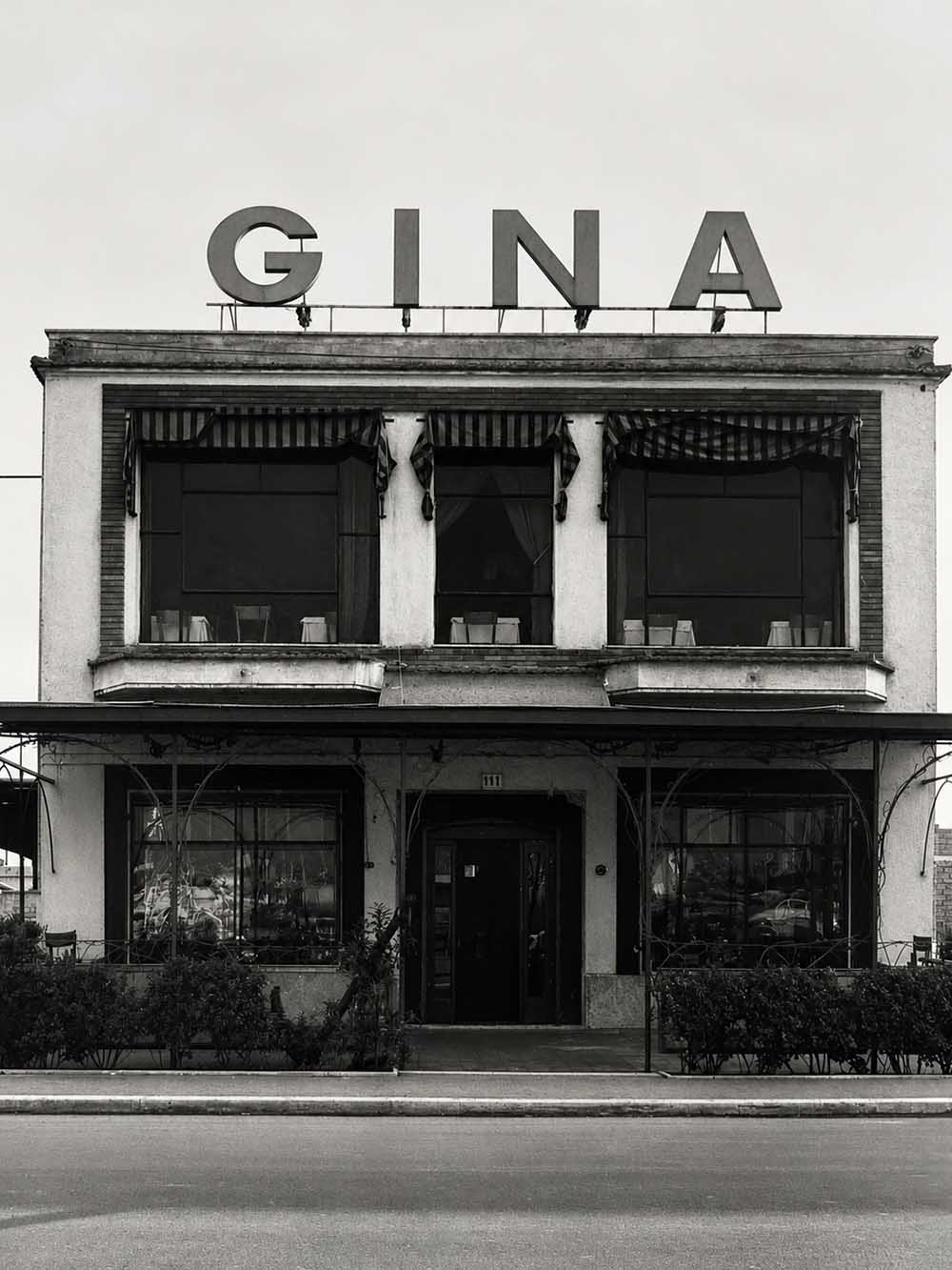 Fotografia in bianco e nero della facciata di Gina Hostaria del Porto: l'insegna GINA a lettere alte sul tetto, le tende a righe sulle finestre del primo piano e l'ingresso al civico 111.