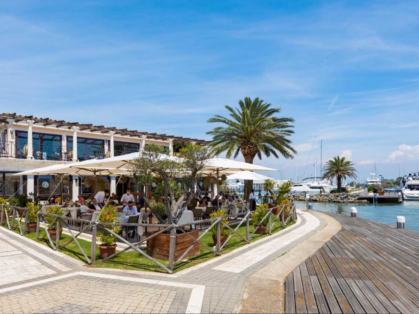 Il ristorante alla Marina Resort di Porto Romano: la terrazza con gli ombrelloni bianchi e i tavoli occupati, sopra la galleria coperta con le colonne bianche e le vetrate, il giardino recintato in legno, la palma e la banchina in legno affacciata sull'acqua con le barche ormeggiate.