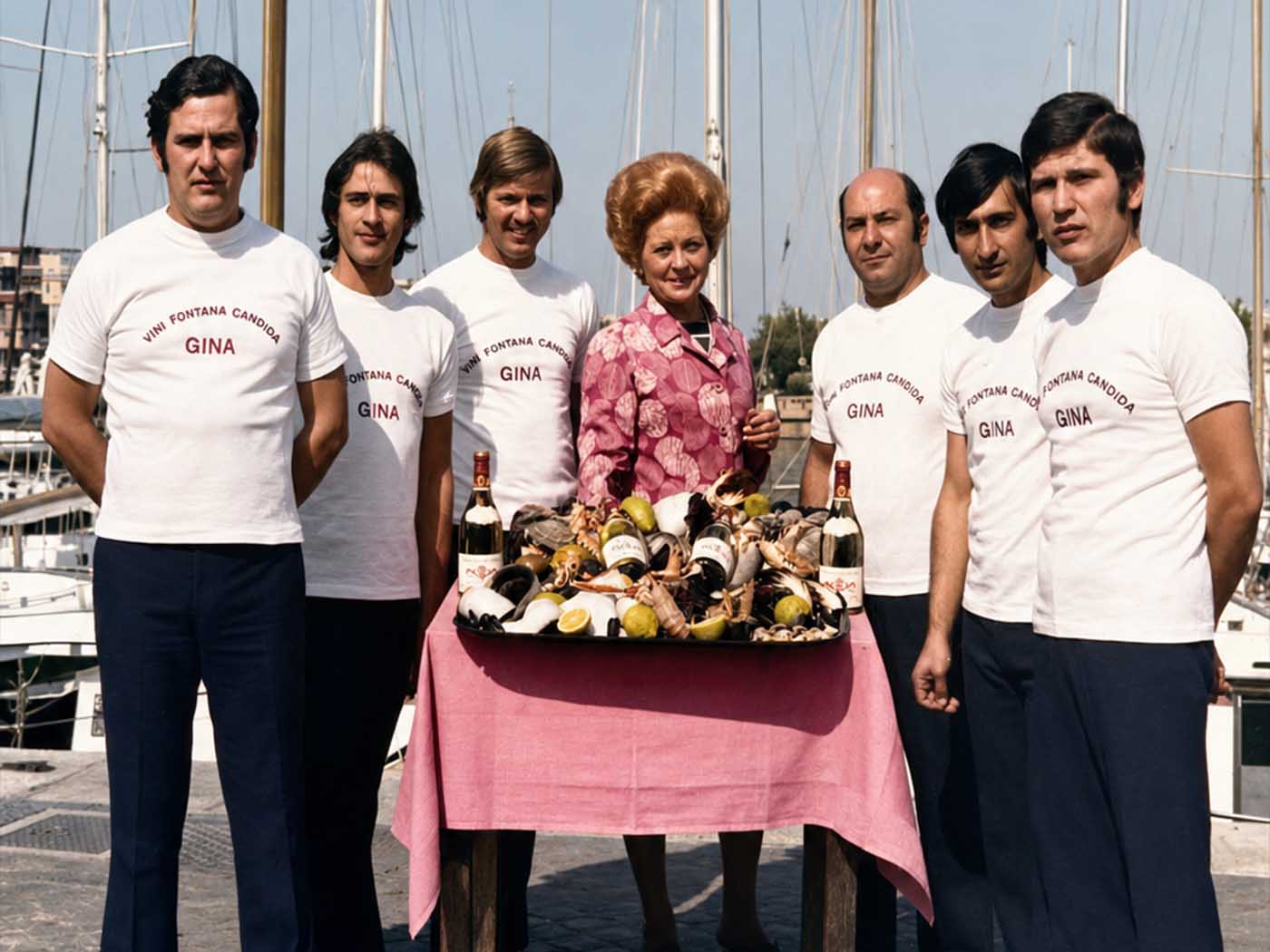 Gina, in giacca rosa, in posa sulla banchina insieme a sei uomini della brigata in maglietta bianca con la scritta GINA; davanti a loro un vassoio di crostacei e frutti di mare e due bottiglie di vino, sullo sfondo le barche ormeggiate.