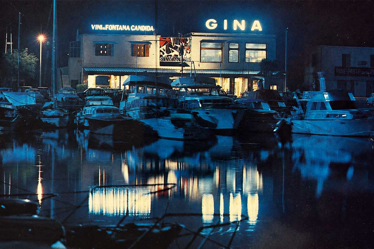 Il ristorante di notte visto dall'acqua della darsena: le insegne luminose GINA e Vini Fontana Candida si riflettono sullo specchio d'acqua tra le barche ormeggiate.